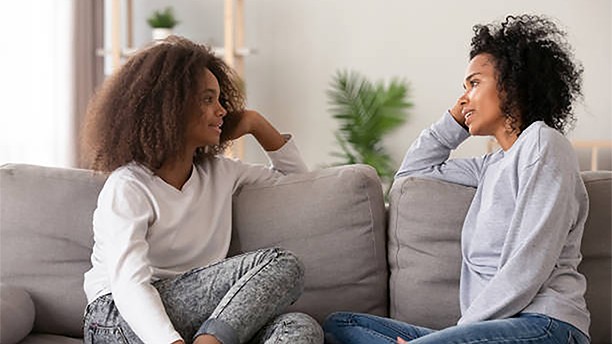 Young african black mother chatting with teen daughter, older sister or friendly nanny and adolescent girl talking sitting together on sofa at home. Good relations different aged people trust concept